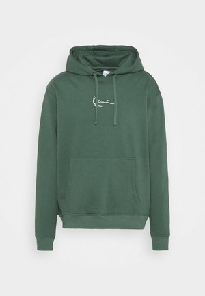 Homme portant un sweat à capuche vert, un sous-pull rouge, un pantalon vert foncé, des chaussettes blanches et des baskets blanches avec des accents verts et noirs, debout contre un mur blanc.