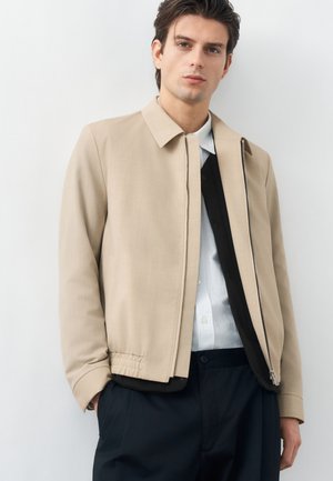 Νεαρός άνδρας που φοράει μπεζ μπουφάν με φερμουάρ πάνω από μαύρο γιλέκο και λευκό πουκάμισο με γιακά, στέκεται μπροστά σε μια απλή ανοιχτόχρωμη επιφάνεια.