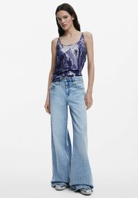 Fialový top na ramienkach s potlačou palmy, skombinovaný so svetlomodrými širokými džínsami. Viditeľná denimová textúra, uvoľnený strih. Tenisky dotvárajú vzhľad.