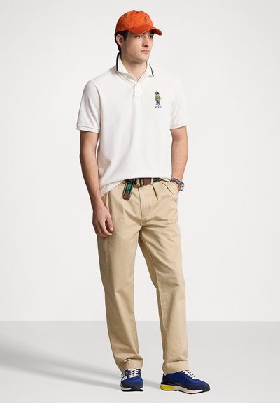 Polo blanc avec col vert, logo brodé ; pantalon kaki ; casquette de baseball orange ; baskets bleues avec accents jaunes ; ceinture multicolore.