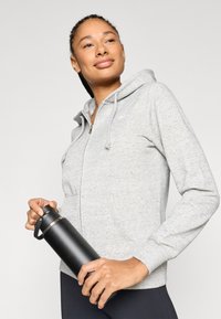 Γυναίκα με γκρι φούτερ με φερμουάρ που κρατάει μια μαύρη μονωμένη φιάλη νερού, κοιτάζοντας ελαφρώς προς τα επάνω και στο πλάι, με φόντο ένα απλό τοπίο.