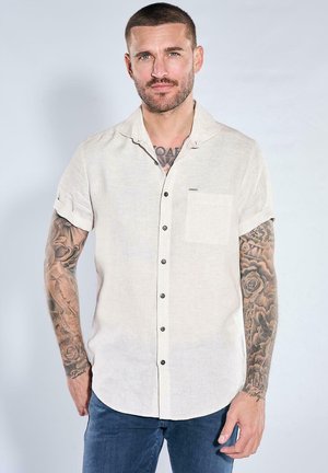 Mann mit kurzen Haaren und Tätowierungen an beiden Armen, der ein beiges kurzärmeliges Hemd und blaue Jeans vor grauem Hintergrund trägt.