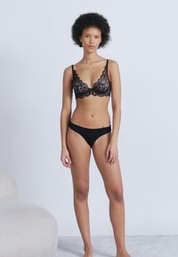 Γυναίκα με σγουρά μαλλιά φορώντας μαύρο δαντελένιο σουτιέν και ταιριαστό μαύρο εσώρουχο, όρθια ξυπόλητη σε φωτεινό χαλί με απλό φόντο.