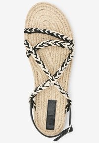 Sandaal in touwstijl met een geweven voetbed van natuurlijk jute, zwart en crème gevlochten banden, en een zwart leren accent met een label.