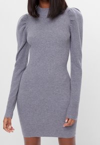 Kvinna klädd i en åtsittande grå långärmad klänning med ribbat mönster, puffärmar och hög halsringning, stående mot en enfärgad bakgrund.