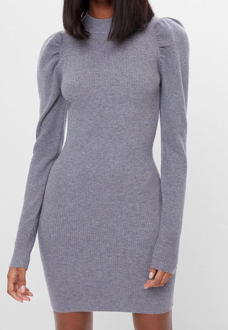 Kvinna klädd i en åtsittande grå långärmad klänning med ribbat mönster, puffärmar och hög halsringning, stående mot en enfärgad bakgrund.