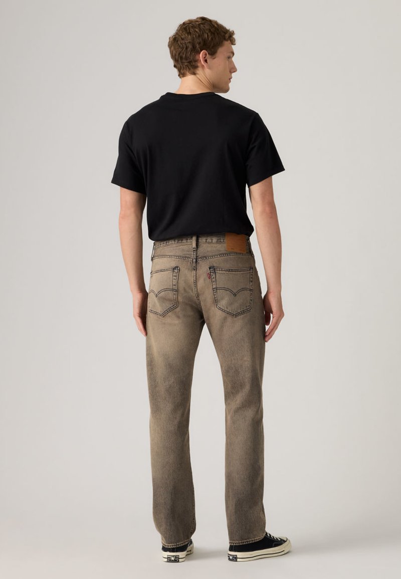 Hombre de pie de espaldas, vistiendo una camiseta negra, jeans marrones desgastados de pierna recta y zapatillas negras y blancas, sobre un fondo liso.