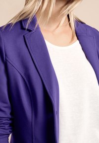 Fioletowa marynarka o strukturalnym kroju, z klapkami i zapięciem na guziki. Noszona na gładkiej białej bluzce, podkreślająca jej wyrazisty kolor.