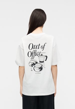 Femme aux longs cheveux foncés portant un T-shirt blanc avec le texte « Out of Office » et des illustrations de tasse de café noir, croissant et tasse au dos.