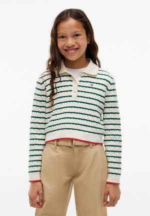 Fille souriante portant un pull à rayures blanches et vertes à manches longues avec un col et un pantalon beige sur fond blanc.