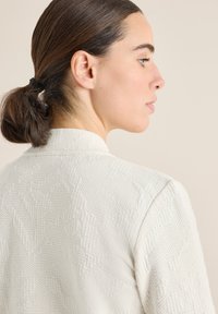 Bílá texturovaná bunda s jemným geometrickým vzorem, s rovný límcem a bezešvými rameny, zobrazená zezadu.