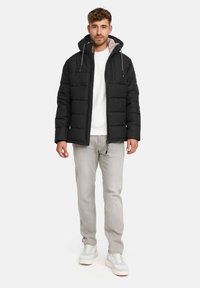 Mann in schwarzer gepolsterter Kapuzenjacke, weißem Sweatshirt, hellgrauen Jeans und weißen Turnschuhen, stehend vor einem weißen Hintergrund.