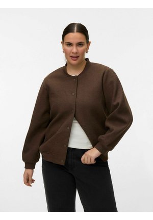 Sötét hajú nő barna gombos zakóban, fehér ingben és fekete nadrágban, egyszerű világos háttér előtt pózol.