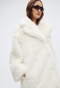 Λευκό παλτό από ψεύτικη γούνα με φαρδιά, μαλακή γιακά και ξεχωριστή, φαρδιά γραμμή. Έχει ομαλή υφή και πλαϊνές τσέπες. Το μοντέλο φορά γυαλιά ηλίου.