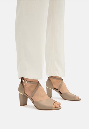 Beżowe buty na obcasie z otwartym palcem, projekt z krzyżującymi się paskami oraz złotym akcentem na blokowym obcasie. Gładka tekstura z minimalnym przeszyciem.