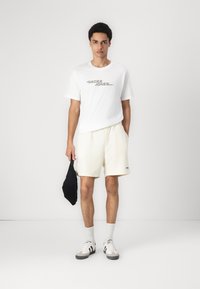 Biała koszulka z szarym logo, jasne kremowe szorty, szaro-czarne sneakersy, białe skarpetki i czarna torba w ręku. Prosty, casualowy strój sportowy.