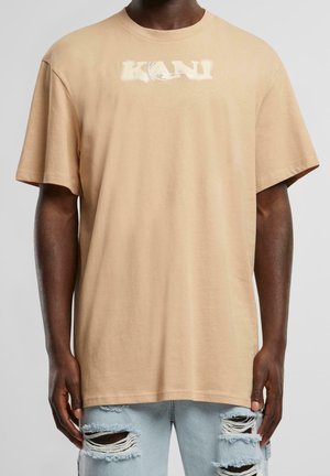 Beżowy oversize t-shirt z wyblakłym logo KANI noszony przez osobę o ciemnej karnacji i podartych jasnoniebieskich dżinsach na szarym tle.