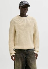 Pull en tricot beige à coupe ample, col rond et poignets côtelés, associé à un pantalon sombre et ample et une casquette grise.