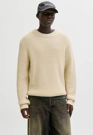 Beige neulepaita, jossa on rento istuvuus, pyöreä pääntie ja resorit hihansuissa, yhdistetty tummaan, väljästi istuvaan housuun ja harmaaseen lippa- tai pipoalueeseen.