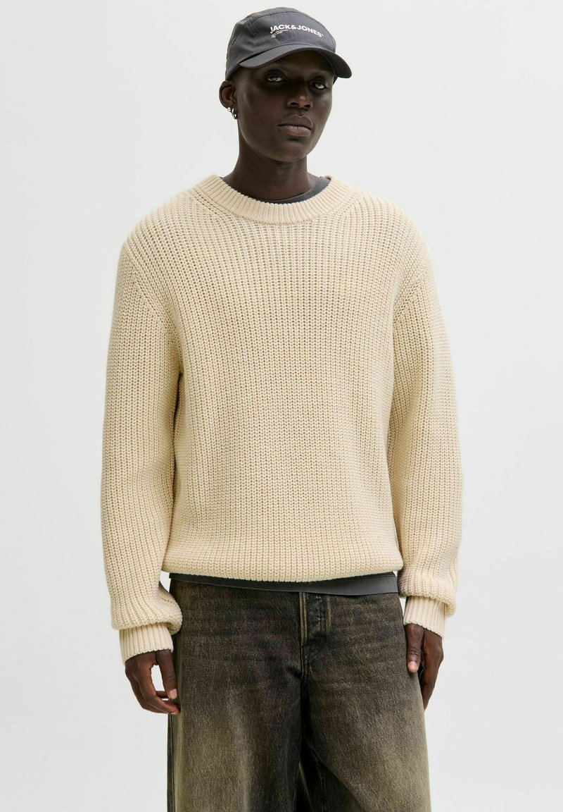 Pull en tricot beige à coupe ample, col rond et poignets côtelés, associé à un pantalon sombre et ample et une casquette grise.