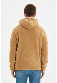 Beżowa bluza z kapturem z miękkiego polaru, o luźnym kroju i kapturze ze sznurkiem. Widok od tyłu ukazuje ściągacze na rękawach i dole bluzy. Noszona z niebieskimi dżinsami.