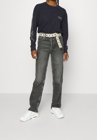 Mörk långärmad skjorta med "FRED" tryckt på ärmen, matchad med högmidjade grå jeans och ett vitt bälte med silverdetaljer.