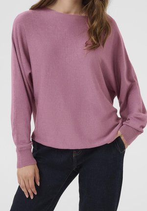 Kvinde iført en løs, langærmet lilla sweater med ribbede ærmekanter og mørkeblå jeans, den ene hånd i lommen, foran en ensfarvet baggrund.