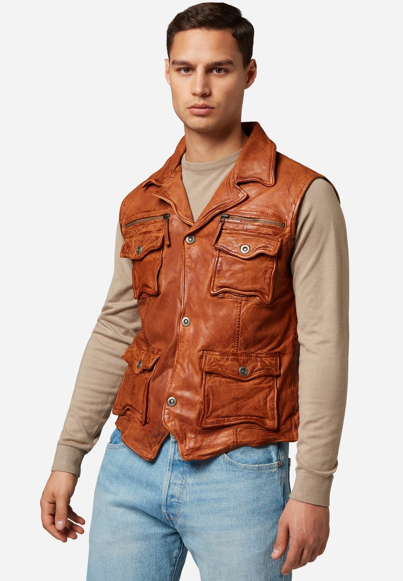 Brauner Lederweste mit mehreren Fronttaschen, einem Kragenhälse und Knopfverschlüssen. Über einem beigen Langarmshirt getragen.