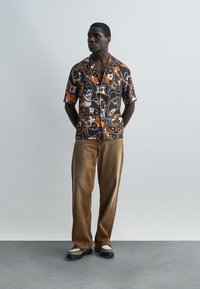 Hombre de pie con las manos detrás de la espalda, vistiendo una camisa marrón de manga corta con estampado floral, pantalones marrones sueltos y zapatos bicolor, frente a una pared gris lisa.