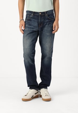 Personne portant un jean en denim bleu foncé et des baskets blanches, debout avec les mains détendues le long du corps sur un fond uni.