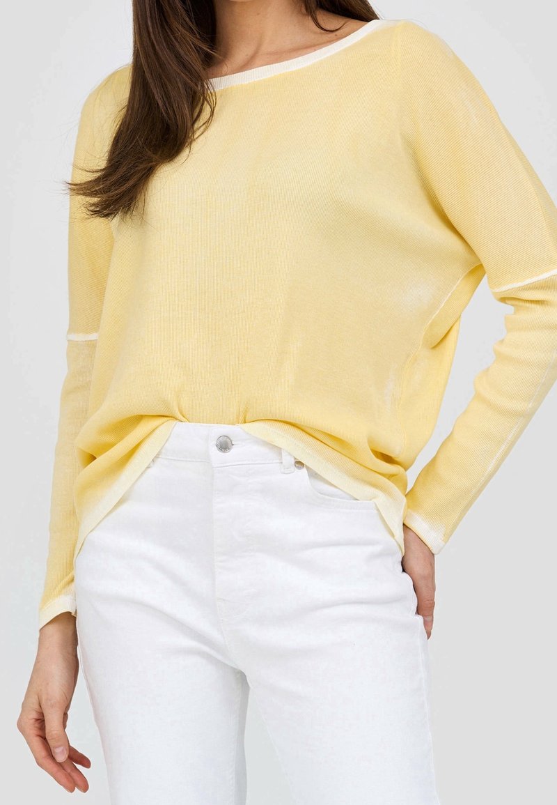 Kevyt, poimullinen villapaita vaaleankeltaisena valkoisilla yksityiskohdilla, leveällä kaula-aukolla ja tiputetuilla hartioilla, yhdistettynä valkoisiin farkkuihin.