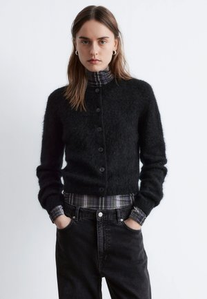 Sort fluffy cardigan med knapper foran, båret over en ternet turtleneck. Cardiganen har lange ærmer og en tætsiddende silhuet.