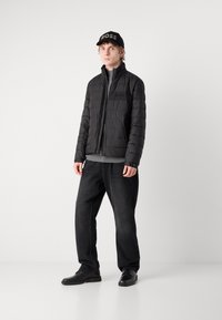 Zwarte pufferjack met ritssluiting, gestructureerd patroon, grijze longsleeve shirt, zwarte relaxed-fit jeans en zwarte schoenen met een platte zool.