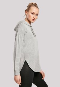 Hellgrauer Kapuzenpullover mit lockerer Passform, seitlichen Schlitzen und gerippten Bündchen. Weiche Textur, kombiniert mit schwarzen Leggings im Hintergrund.