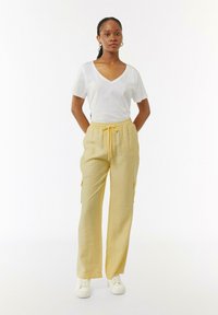 Weißes V-Ausschnitt-T-Shirt und gelbe, weiche, leichtgewichtige Cargo-Hosen mit elastischem Bund und zwei Seitentaschen. Weiße Sneaker runden das Outfit ab.