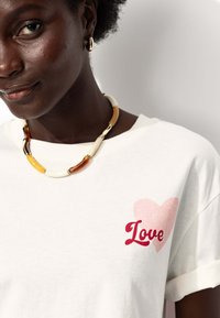 T-shirt en coton blanc avec un graphique en forme de cœur rose et le mot "Love" en rouge. Le modèle porte un collier de perles multicolores.