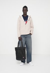 Juodas rankinis krepšys su prekiniu ženklu diržais, priekinis kišenė, derinamas su kreminių spalvų švarku, tamsiai mėlynu grafiniu marškinėliu ir šviesiai mėlynomis džinsinėmis kelnėmis.