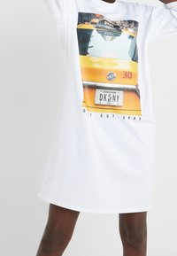 Weißes Baumwoll-T-Shirt-Kleid mit einem Grafikdruck eines gelben Taxis, einschließlich des Nummernschilds "DKNY" und dem Text "NEW YORK" darunter.