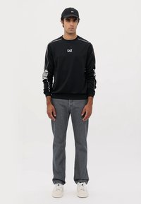 Felpa nera con dettagli bianchi, presenta il logo EA7 e un design a righe sulle maniche. Abbinata a jeans grigi e sneakers bianche.