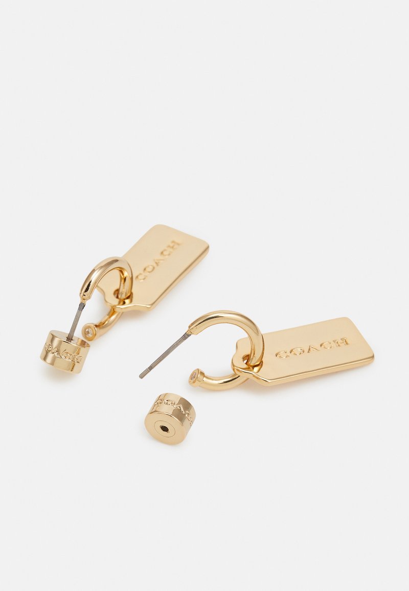 Boucles d'oreilles en or avec un design de tag rectangulaire. Présentent le nom de la marque gravé et des accents cylindriques. Texture lisse avec une finition brillante.