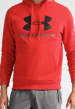 Czerwona bluza z kapturem Under Armour z czarnym logo i napisem, noszona ze szarymi spodenkami dresowymi, pokazująca górną część ciała i dłonie.