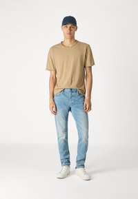 Mlad moški v temno modri kapi, bež majici s kratkimi rokavi, svetlih modrih kavbojkah in belih supergah stoji ob brezbarvni beli ozadju.