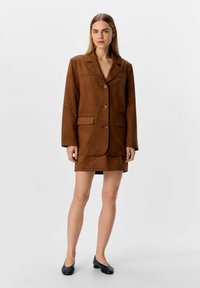 Blazer en daim marron avec revers crantés, trois boutons et poches avant, associé à une jupe assortie et des escarpins noirs. Texture lisse.