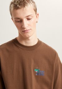 Bruine katoenen sweatshirt met een ronde hals, korte mouwen en een kleurrijke kikkerafbeelding met tekst op de borst. Zachte textuur.