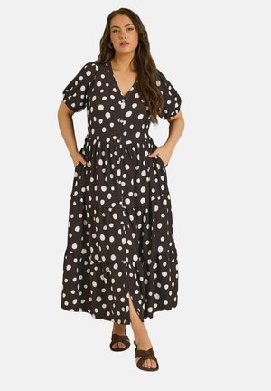 Kvinde iført en sort midi-kjole med hvide polkaprikkker, korte pufærmer, knapper foran og brune sandaler, stående med hænderne i lommerne.