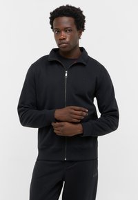 Felpa nera con zip e colletto alto, realizzata in tessuto morbido, con un logo discreto sul petto e polsini e orlo a coste.