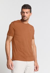 Korte bruine t-shirt met korte mouwen van een lichtgewicht, soepel materiaal met een ronde hals. Kleine logo detail op de borst. Model draagt lichtgekleurde broek.