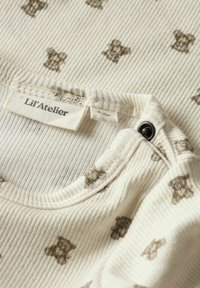 Krämfärgad ribbad tyg med nalles mönster, med rund halsringning och tryckknappsstängning. Etiketten säger "Lil' Atelier."