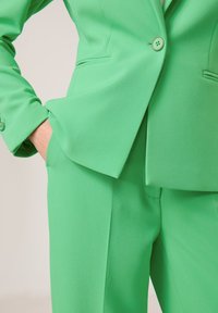 Grüner Anzug mit einem taillierten Jackett mit einem einzigen Knopf und weit geschnittenen Hosen. Das Material ist glatt mit matter Oberfläche.