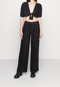 Top noir cropped avec un devant noué et de courtes manches bouffantes, associé à un pantalon large taille haute noir avec des coutures blanches.